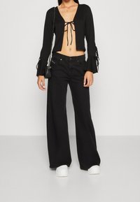 Sort cropped bluse med bindingslukning og flagrende ærmer, parret med brede sorte jeans og hvide platform sneakers. Minimalistisk sort taske.