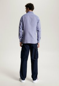 Camicia a maniche lunghe a quadri blu con colletto, abbinata a pantaloni cargo blu navy con molteplici tasche e sneakers bianche con dettagli rossi.