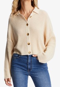 Stickad cremefärgad cardigan med krage, knäppning fram och texturerat mönster. Matchas med högmidjade blå jeans.