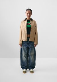 Beige cotton jacket with a brown collar, four pockets, worn over a black graphic t-shirt. Paired with loose, faded blue denim jeans and green sneakers.