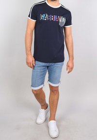 Námořnické modré tričko s vícebarevným nápisem "GABBIANO"; bílé akcenty na rukávech a krku. Spárováno s ohrnutými džínovými šortkami a bílými teniskami.