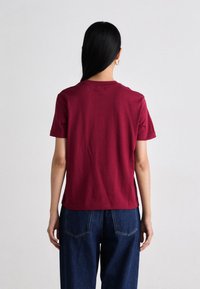 Personne avec de longs cheveux noirs et des boucles d'oreilles en or, portant un simple t-shirt bordeaux à manches courtes et un jean bleu, de dos sur un fond blanc.