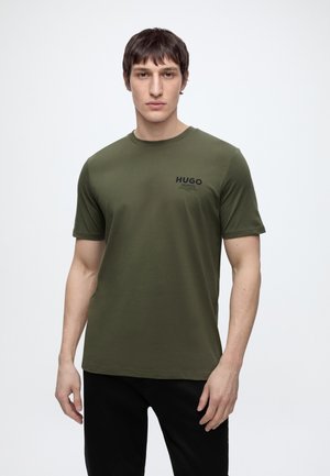 Ung mand iført en olivengrøn Hugo t-shirt og sorte bukser, der står mod en ensartet hvid baggrund.