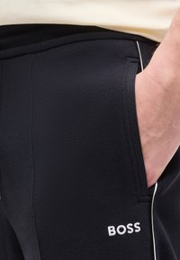 Χέρι ατόμου που βρίσκεται στην τσέπη μαύρου υφασμάτινου παντελονιού με λευκό λογότυπο "BOSS" και λευκή επένδυση κατά μήκος της ραφής.