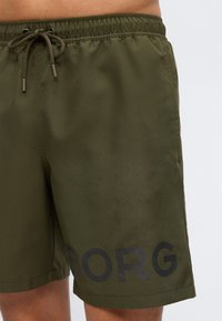 Ανδρική βερμούδα σε ελιά πράσινο με ελαστική μέση και κορδόνι, με μεγάλο μαύρο κείμενο «BORG» κοντά στο τελείωμα του αριστερού ποδιού.