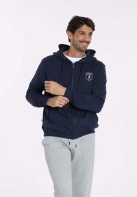 Sweat à capuche zippé bleu marine avec cordon de serrage, orné d'un patch logo sur la poitrine, porté avec un pantalon de survêtement gris. Texture du tissu douce.