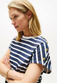 Camiseta de rayas en azul marino y blanco con mangas cortas, cuenta con acentos de botones dorados en el hombro y un cuello redondo. Textura de tela suave.