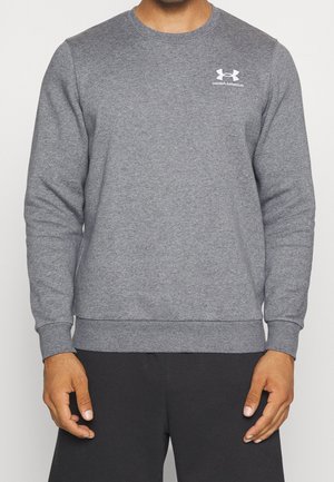 Mężczyzna ubrany w prostą szarą bluzę z długim rękawem i okrągłym dekoltem, z małym białym logo Under Armour na lewej piersi oraz czarne spodnie.