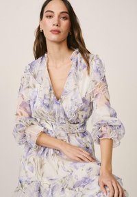 Mujer con un vestido transparente con volantes y estampado floral en tonos suaves de púrpura y verde, con pendientes dorados de aro, sentada frente a un fondo liso.