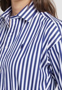 Kvinna som bär en blå och vit vertikalt randig skjorta med knappar och en polo-spelare broderad på bröstet.