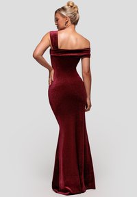 Vrouw met blond haar in een hoge knot, gekleed in een aansluitende, een schouder bloot latende, fonkelende donkerrode avondjurk, staand met haar rug naar de kijker.