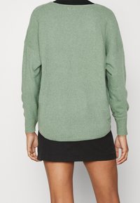 Maglione verde a maglia con spalle scese, scollatura a girocollo e polsini a costine, abbinato a una gonna nera. Tessuto morbido e vestibilità rilassata.