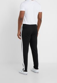 Man som bär svarta träningsoverallbyxor med vita sidostygn, vit t-shirt och vita sneakers som står mot en enfärgad bakgrund.