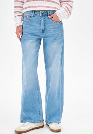 Osoba ubrana w jasnoniebieskie jeansy o szerokich nogawkach, białe trampki i różowo-biały sweter w paski, stojąca z jedną ręką w kieszeni.