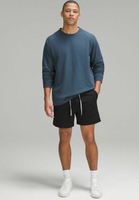Sweat-shirt à manches longues de couleur bleu sarcelle foncé avec des bords côtes, associé à un short noir. Le modèle porte des baskets blanches et des chaussettes de cheville.