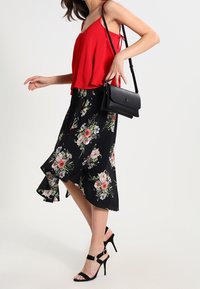 Kvinna som bär en röd ärmlös topp, svart blomster midi-kjol, svarta högklackade sandaler och bär en svart axelväska.