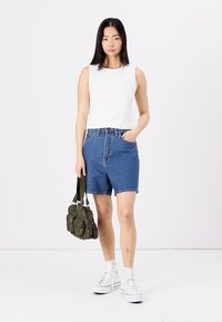 Top blanc sans manches, short en denim bleu taille haute, baskets blanches à plateforme, sac à main vert avec plusieurs poches, debout sur un fond uni.