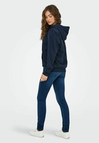 Sweatshirt à capuche navy avec une coupe décontractée, manches côtelées, associé à un jean bleu ajusté et des baskets blanches. Fond blanc uni.