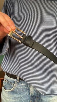 Črn usnjen pas z zlatim kovinskim sponko, ki kaže gladko teksturo. Nošen čez modro in belo črtasto srajco z džins hlačami.