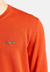 Maglione lavorato a maglia arancione con scollo a V e logo di ali dorato ricamato sul lato sinistro del petto. Tessuto texturizzato e cuciture dettagliate sulla spalla.