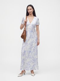 Femme portant une robe longue blanche et bleu clair à motifs floraux avec des manches courtes, tenant un sac à bandoulière marron et portant des chaussures blanches à talons.