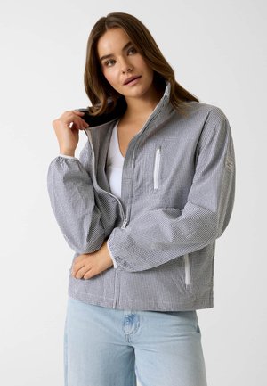 Frau mit braunen Haaren, die hellblaue Jeans und eine graue karierte Zipperjacke trägt, während sie den Kragen richtet.