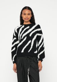 Femeie îmbrăcată într-un pulover în dungi alb-negre cu model zebra și pantaloni negri cu dungi subțiri, cu o centură cu mărgele, stând pe un fundal alb.