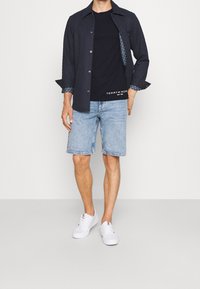 Muž v námořnické prošívané bundě, pod kterou má černé tričko Tommy Hilfiger, světle modré džínové šortky a bílé tenisky, na jednobarevném pozadí.