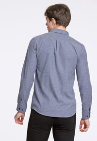 Niebieska koszula z długim rękawem o fakturowanej tkaninie, z wyraźnym kołnierzem i zapięciem na guziki; widok z tyłu z zaokrąglonym dołem.