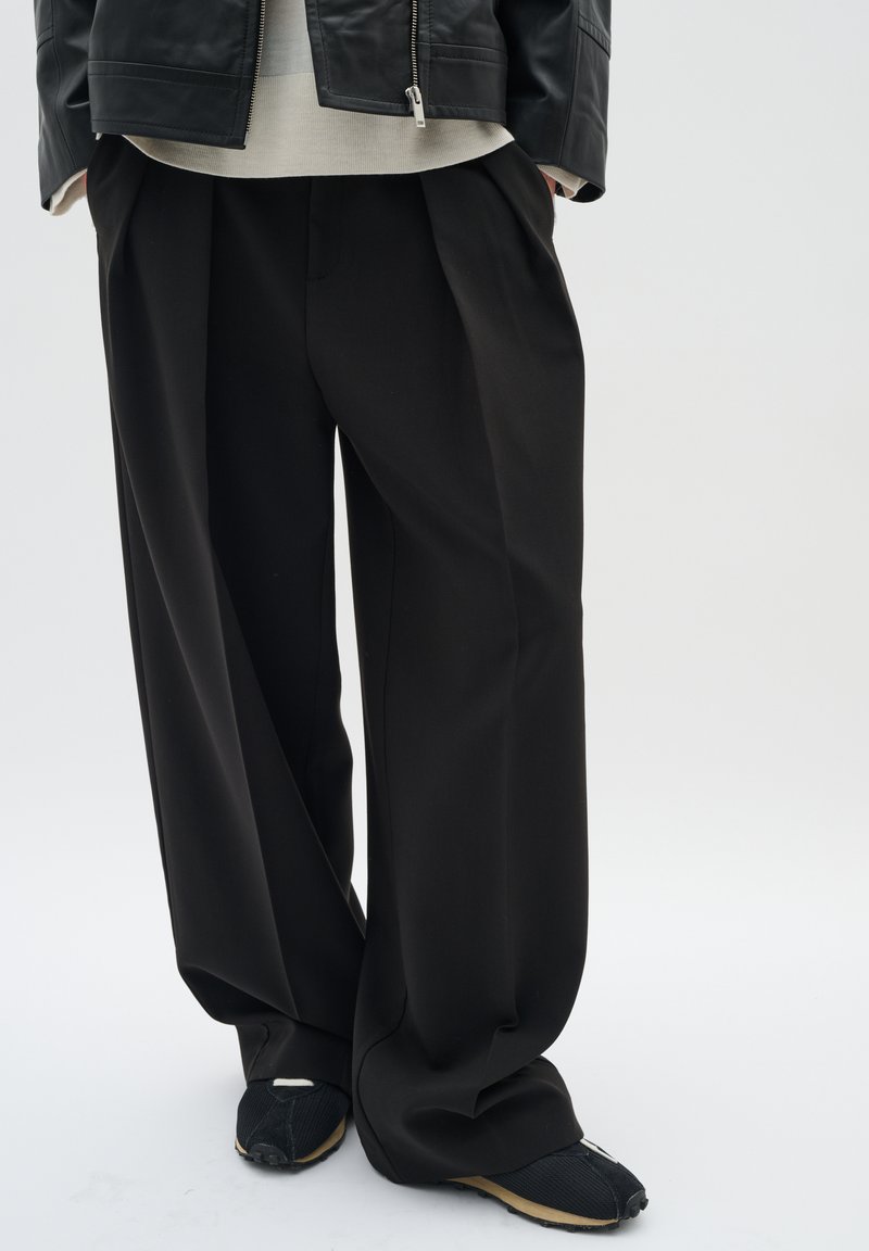 Pantaloni neri a gamba larga con una texture liscia e pieghe frontali, abbinati a scarpe nere con un design testurizzato.