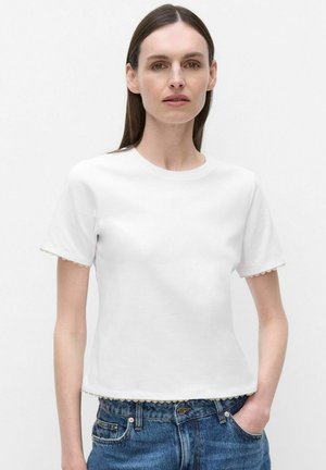 Femme aux longs cheveux bruns portant un haut blanc uni à manches courtes avec des bords festonnés et un jean bleu, debout sur un fond blanc.