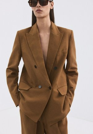 Femme portant un blazer marron croisé et un pantalon assorti, les mains dans les poches et portant de grandes lunettes de soleil foncées.