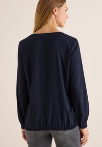 Blouse manches longues bleu marine avec une coupe décontractée, ourlet élastiqué et poignets boutonnés, fabriquée en tissu lisse. Design simple sans motifs visibles.
