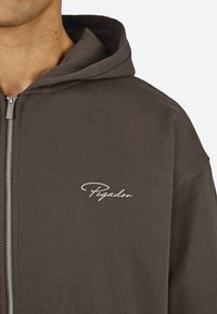 M�ørkegrå zip-up hettegenser laget av myk stoff. Har en frontglidelås og en liten hvit "Pegador"-logo på brystet.