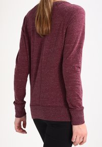 Bordová dlouhá rukávová košile s mírně texturovanou tkaninou, kulatým výstřihem a žebrovanými manžetami, která má uvolněný střih a detail švu na zadní straně.