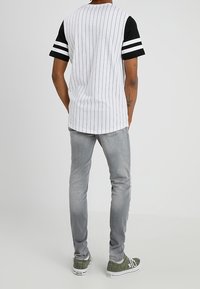 Persona con una camisa blanca a rayas con mangas negras, jeans gris claro y zapatillas verdes, de pie de espaldas en un fondo liso.
