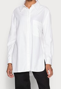 Chemise blanche à manches longues, en tissu de coton lisse, avec une poche poitrine et des fentes sur les côtés, dotée d'un col classique.