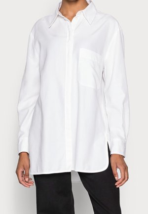 Valkoinen pitkähihainen nappipaita, joka on valmistettu sileästä puuvillakankaasta. Paidassa on rintatasku ja sivuhalkiot, ja sen muotoilu on klassinen kauluksellinen.