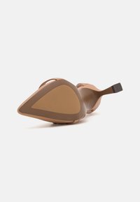 Zapato de tacón alto color beige con punta afilada, correa curvada y suela marrón contrastante. Textura suave con un diseño minimalista.