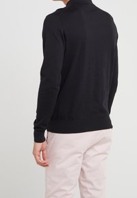 Czarny dziergany sweter z wysokim kołnierzem, dopasowany krój i ściągacze przy rękawach. Noszony z jasnoróżowymi spodniami, widok od tyłu, gładka faktura.