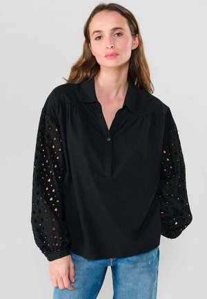 Femme portant un chemisier noir avec des manches bouffantes transparentes à motifs et un jean bleu, se tenant devant un fond gris clair uni.