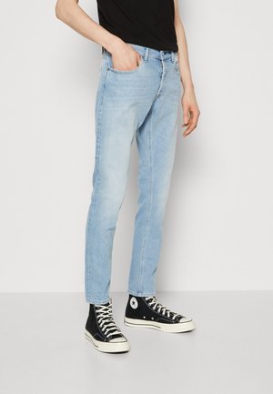 Person iført lyseblå slim-fit jeans, sorte høje sneakers med hvide snørebånd og en sort skjorte med den ene hånd i lommen.