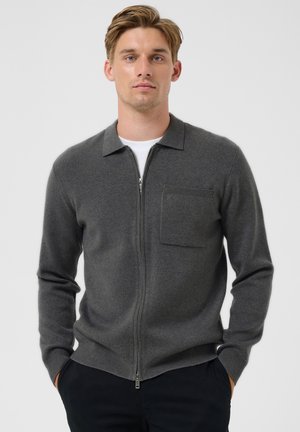 Jeune homme aux cheveux courts portant un pull gris foncé à fermeture éclair sur une chemise blanche, debout les mains dans les poches devant un fond clair.