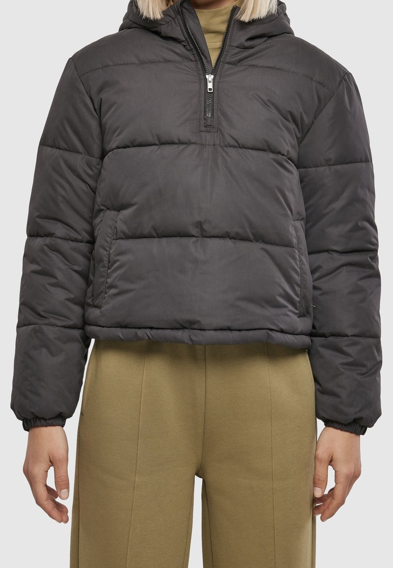 Personne portant une veste matelassée gris foncé à demi-fermeture éclair avec des poignets élastiques et un pantalon taille haute vert olive.