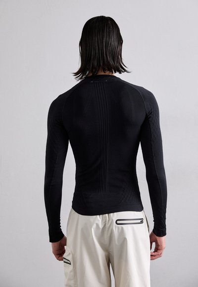 Camisa de desempenho de mangas longas, ajustada ao corpo, na cor preta com padrões texturizados, com detalhes canelados nas costas, acompanhada de calças de tom claro.