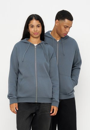 Two people wearing blue zip-up hoodies, one facing forward with hands visible, the other looking down with hands in pockets.