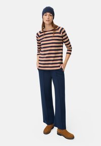 Kvinde iført en marineblå hue, stribet beige og marineblå bluse med trekvartærmer, marineblå vide bukser og brune ruskindsstøvler, stående med hænderne i lommerne.