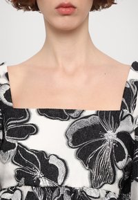 Femme portant une robe à encolure carrée avec une grande broderie florale noire et blanche sur tissu texturé.