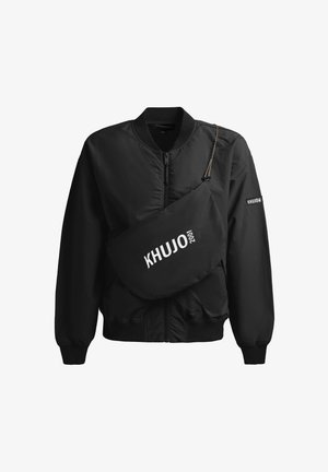 Czarna kurtka typu bomber na zamek z prążkowanymi mankietami i kołnierzem, z czarną torbą na ramię z napisem "KHUJO 2001" w białym kolorze.