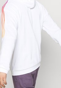 Persoon in een witte hoodie met rood-oranje-geel gestreepte mouwen en paarse cargobroek, arm zijwaarts gestoken.